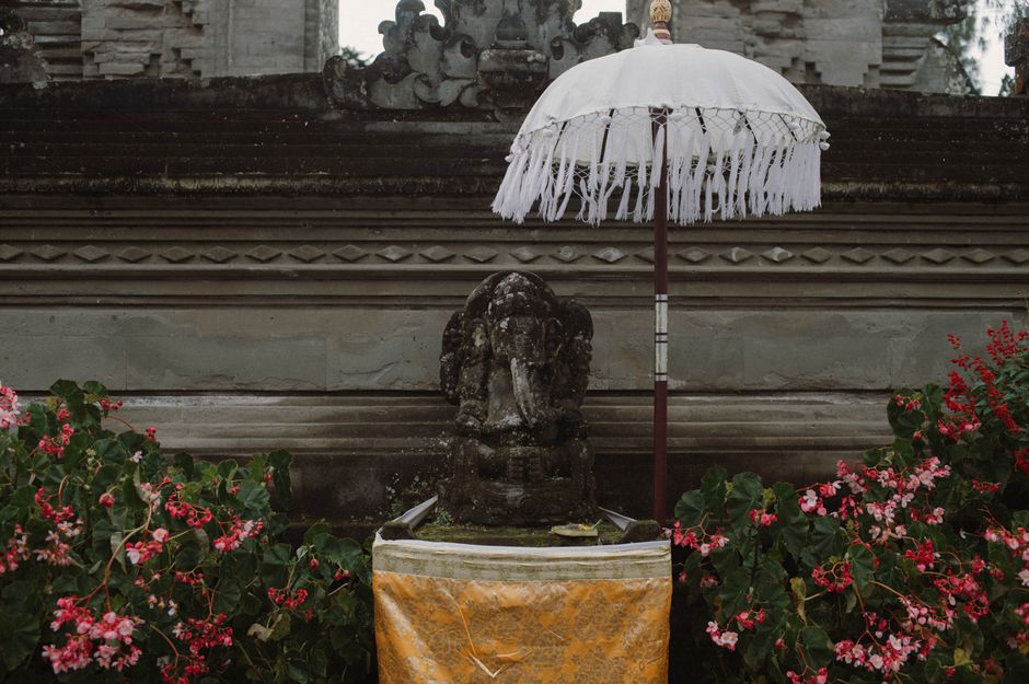 A Balinese stone statue with a white umbrella surrounded by pink flowers in a temple setting.