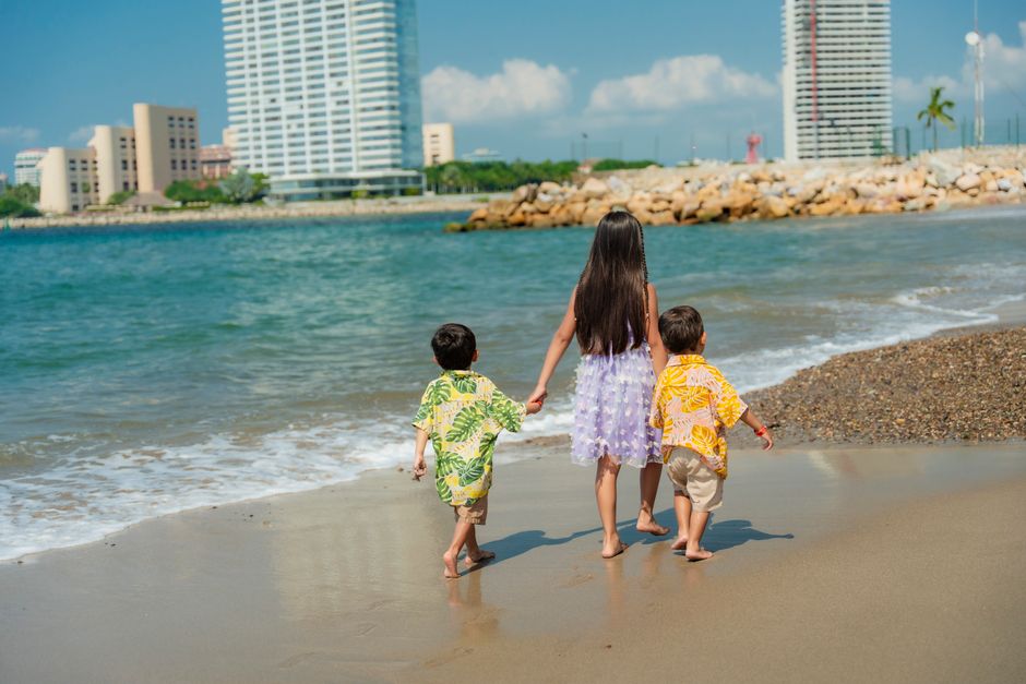 Three kids stroll on a beach, enjoying a sunny day with city skyline backdrop, creating joyful summer memories.