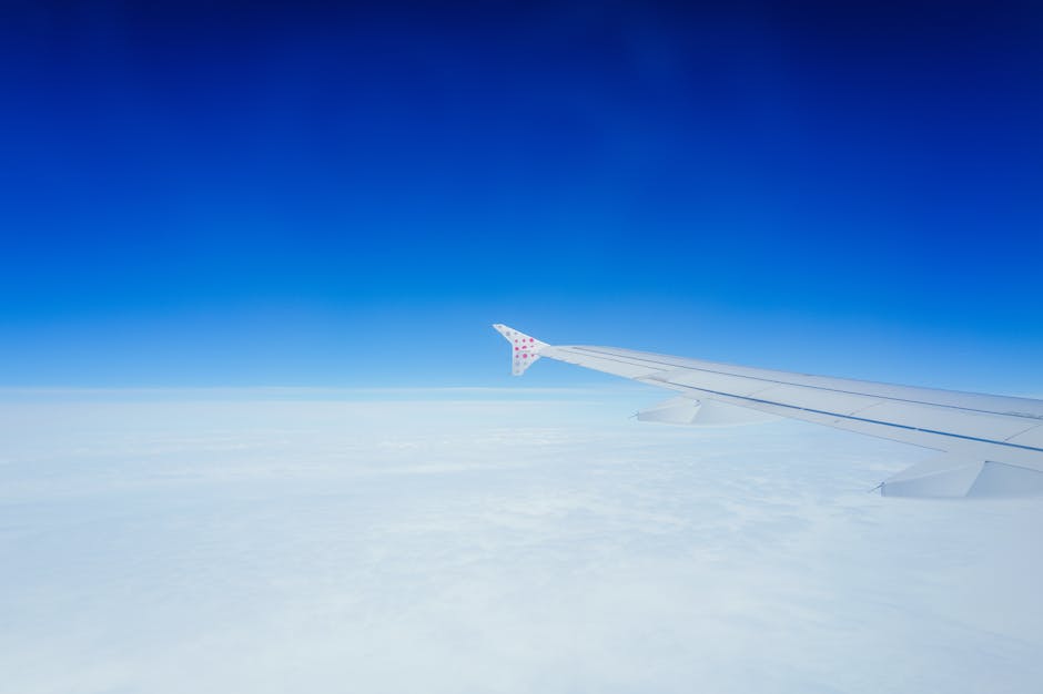 Wing of an airplane against a clear blue sky and fluffy clouds seen from the window.