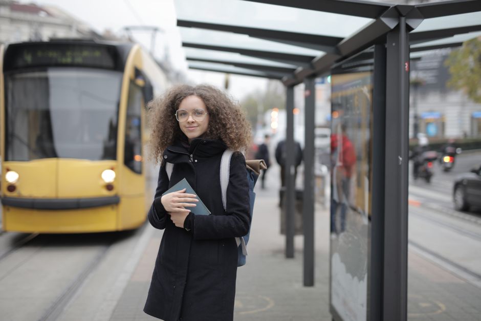 A young woman standing at a tram station holding a book, ready to commute in the city.