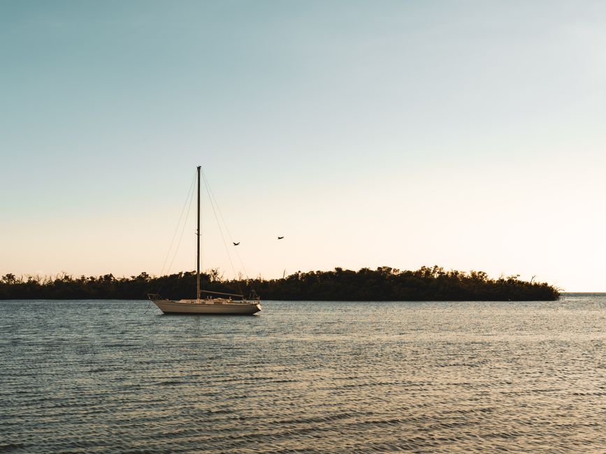 A serene image of a sailboat at sea during sunset, with a clear sky and distant coastline.