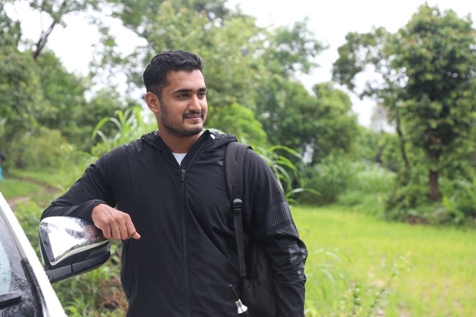 Man in a black jacket leaning on a car outdoors, surrounded by lush greenery.