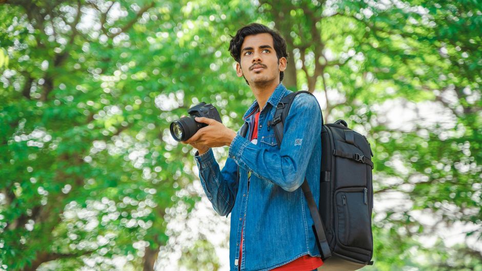 Young man with backpack and camera outdoors capturing photos in a verdant park.