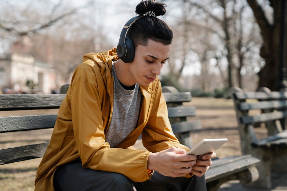 Young ethnic male in casual clothes using tablet and earphones while sitting on bench in park in autumn