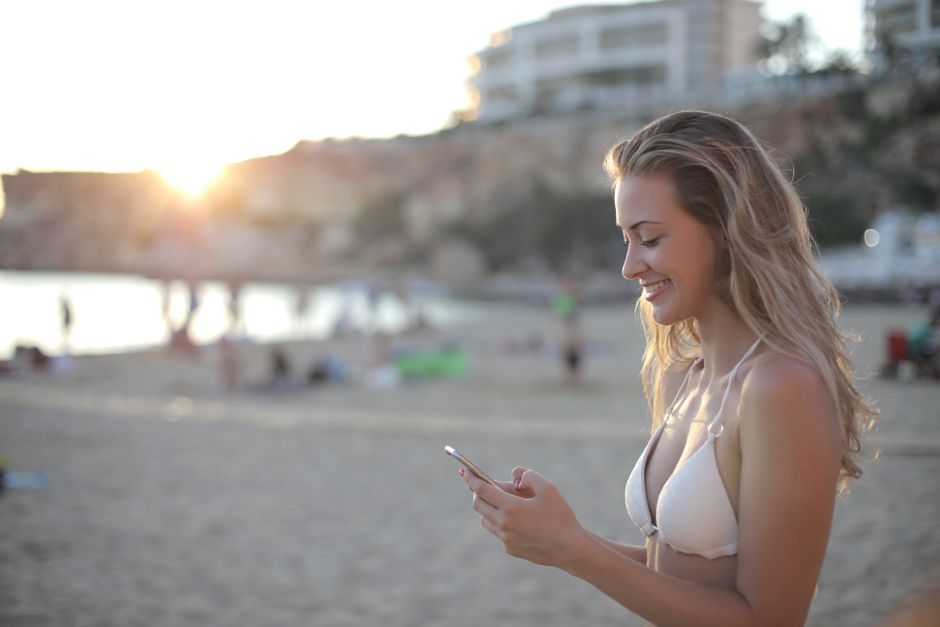 A young woman in a bikini smiles while texting on a sunny beach during sunset.