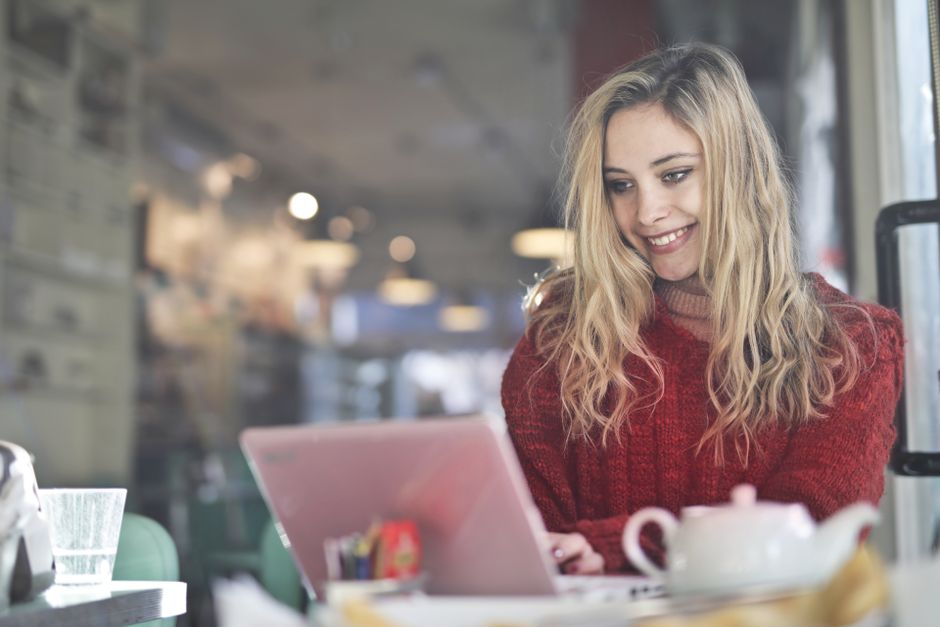A young woman with blonde hair works on a laptop in a cozy cafe, surrounded by a warm ambiance.