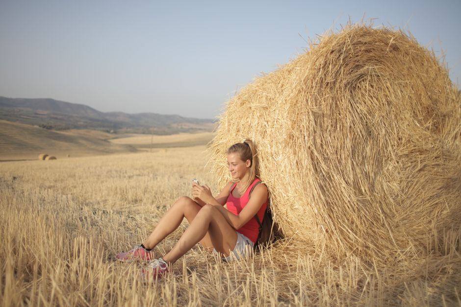 Young woman sitting by a hay bale, enjoying leisure time in a sunny rural field.