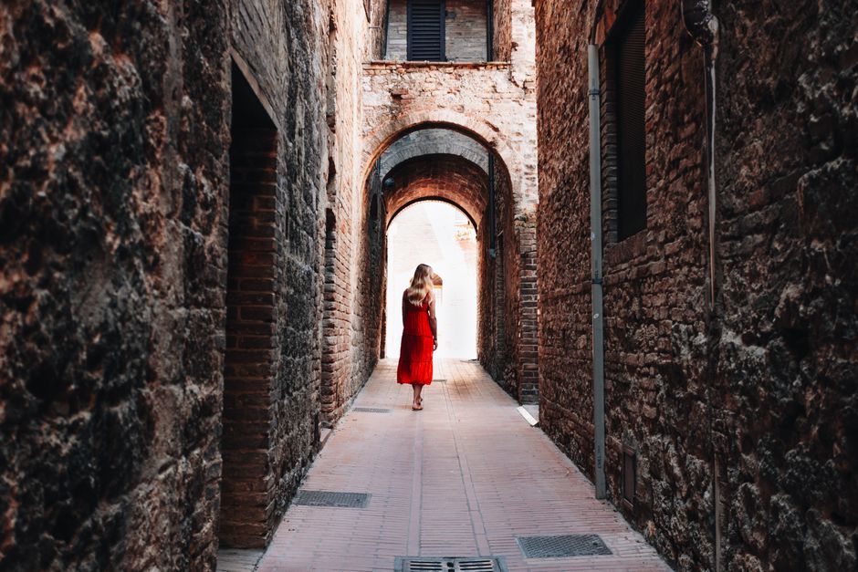 A woman in a red dress walks down a narrow, historic alley lined with brick walls under arches.