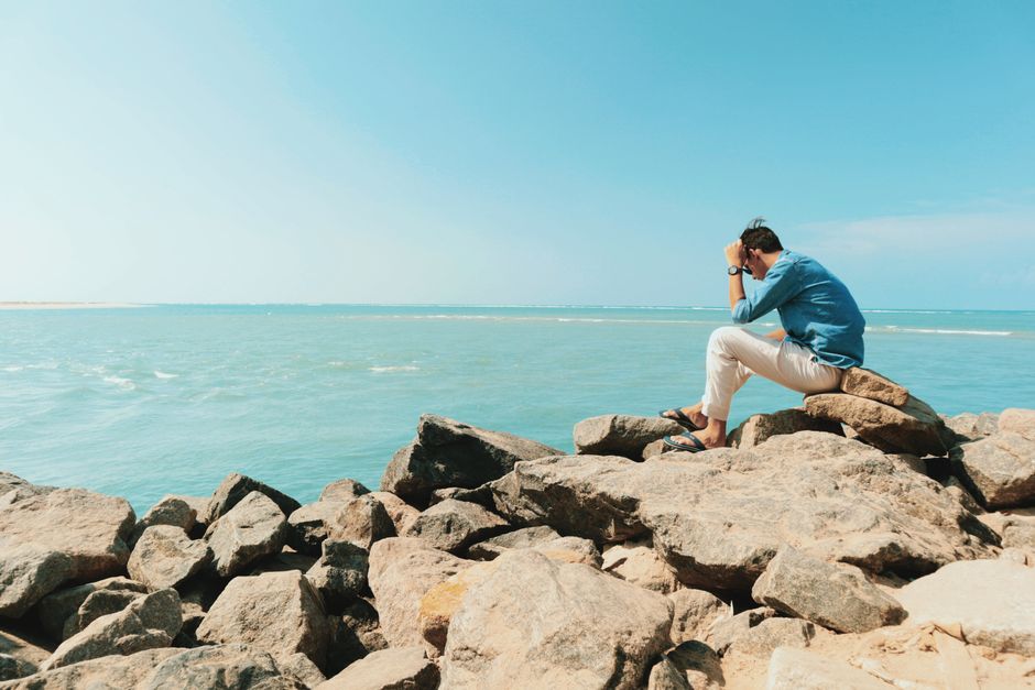 A man in casual attire sitting on rocks by the ocean, enjoying a summer day.