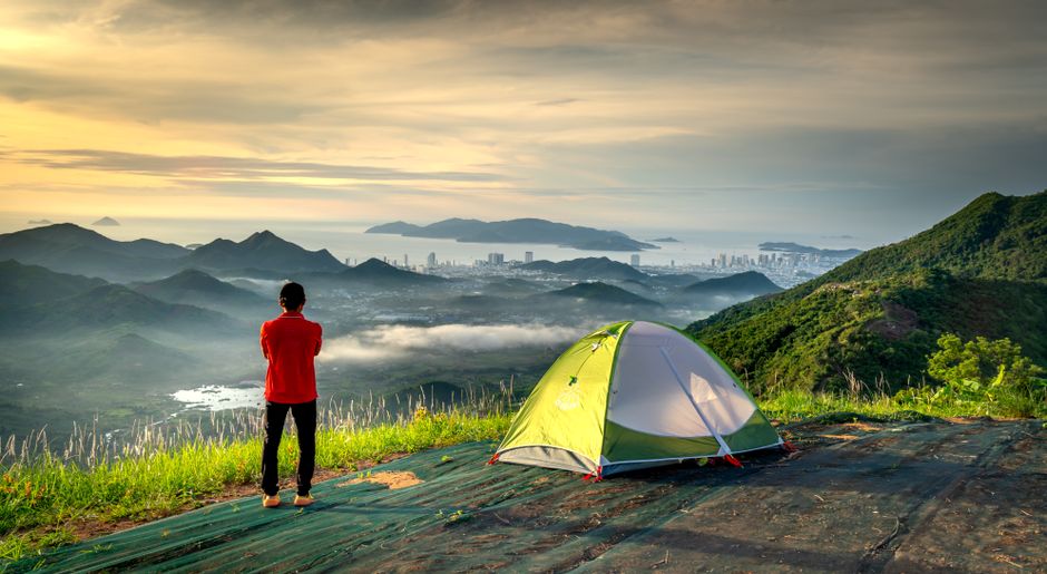 Adventurer stands by tent, embracing scenic mountain and city view at sunrise.
