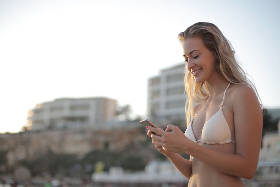 A young woman in a bikini smiles while texting on her smartphone at the beach during summer.